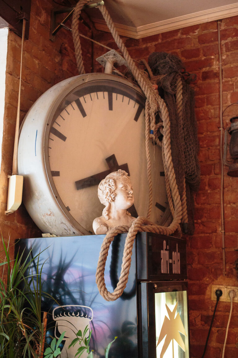 auf eine hohen Getränkekühlschrank liegt eine sehr große Bahnhofsuhr mit Schiffahrtseilen davor ist ist ein antike Büste