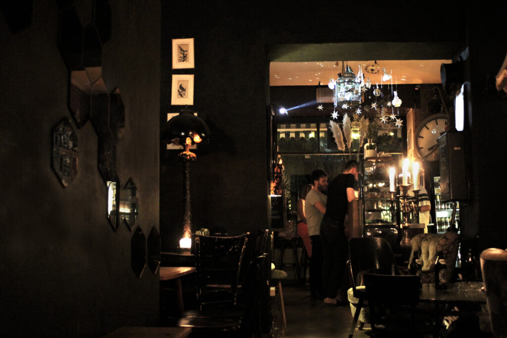 Sicht vom dunklen Raum zur Bar - es stehen 2 Personen an der Bar