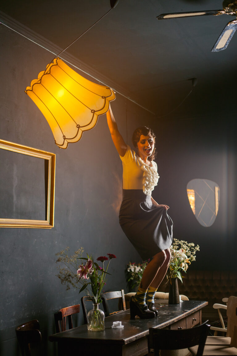 eine Frau tanzt auf einem langen Tisch und schwingt eine bleuchtete Deckenbeleuchtung mit grossen Lampenschirm