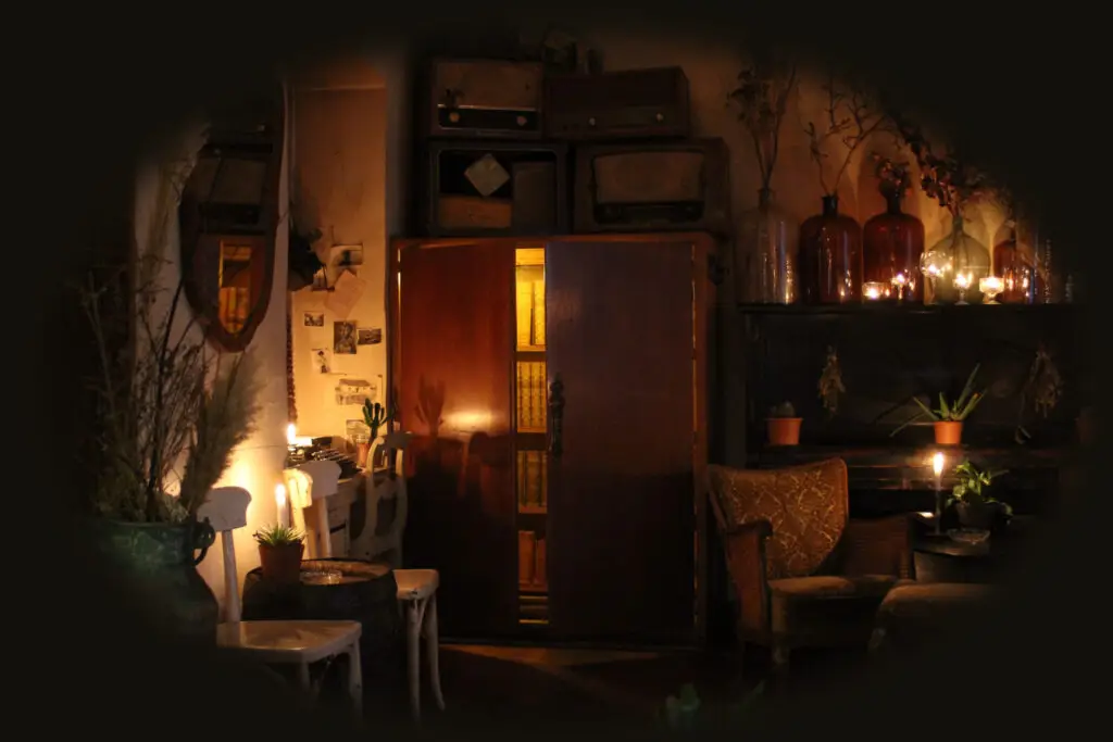 Ein brauner 2 türiger teilweise offener von innen beleuchteter Kleiderkasten. Auf dem Schrank sind antike und alte Radios und Lautsprecher - daneben ist ein schwarzes Piano - es ist der Zugang zu dem geheimen Partyraum